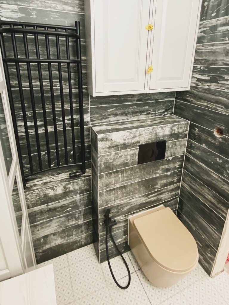 Nowoczesna łazienka z beżową toaletą podwieszaną, czarnym grzejnikiem drabinkowym i szafką wiszącą na tle płytek imitujących drewno.