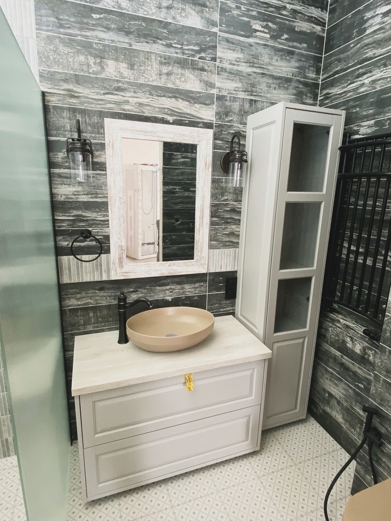 Stylowa łazienka z szarymi płytkami imitującymi drewno i beżową umywalką na szafce w kolorze złamanej bieli. Lustro w ozdobnej ramie, lampy w stylu industrialnym.