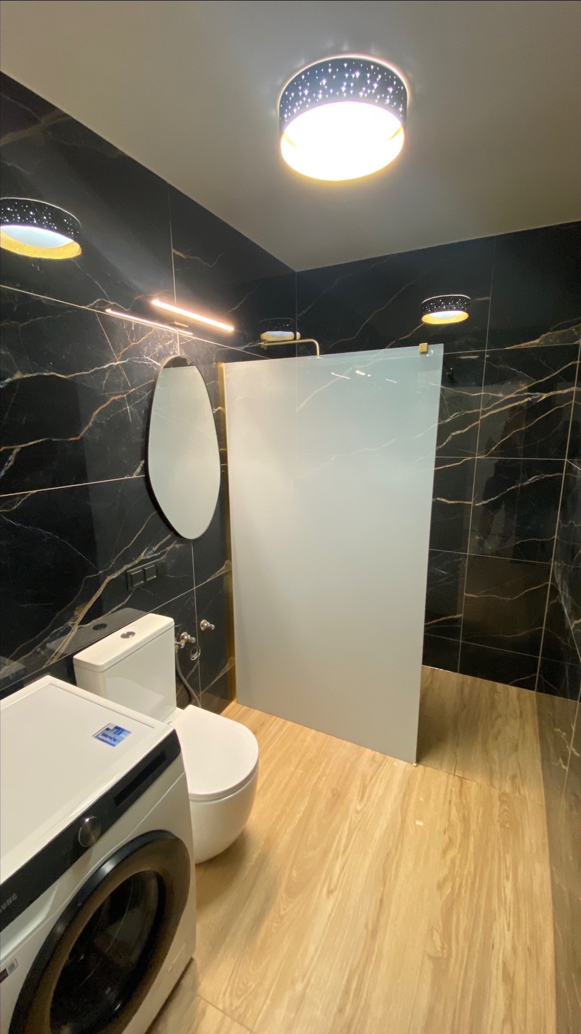 Elegancka łazienka z czarnymi płytkami imitującymi marmur, kabiną walk-in, owalnym lustrem i drewnopodobną podłogą. Widoczna pralka.