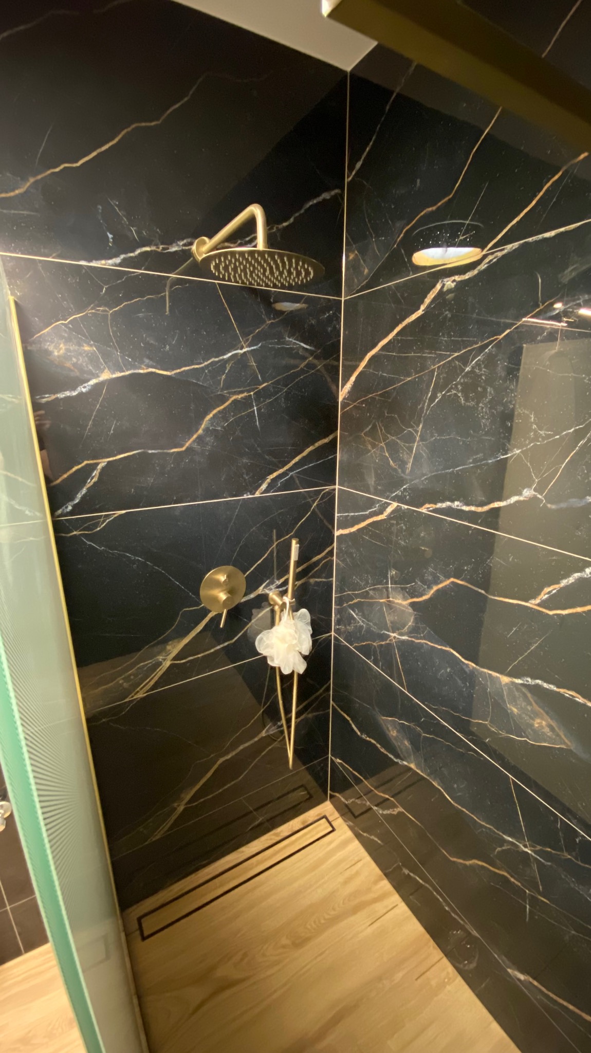 Elegancka kabina prysznicowa z czarnymi płytkami imitującymi marmur, złotą armaturą i odpływem liniowym. Widok na luksusowe wykończenie wnętrza łazienki.