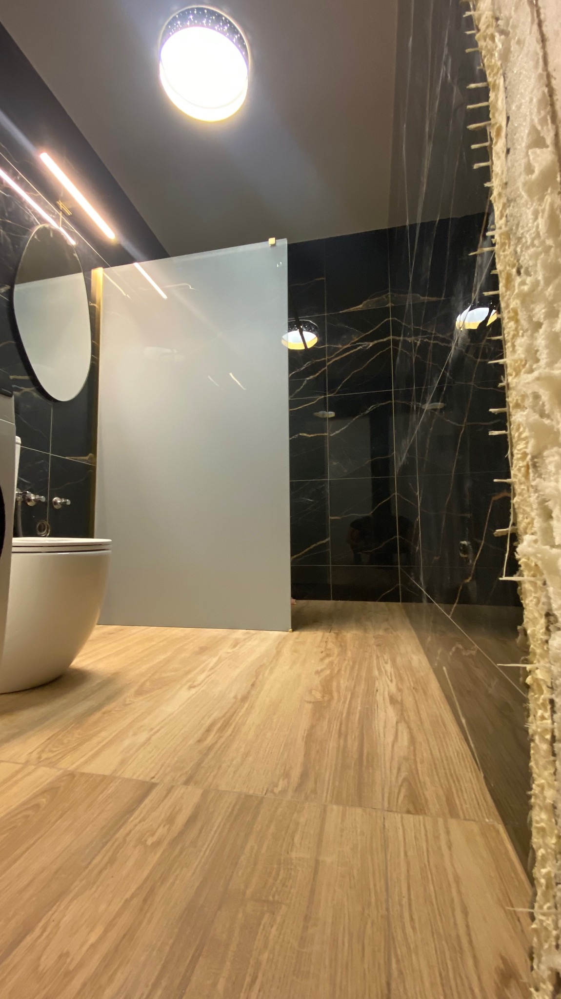 Nowoczesna łazienka z czarnymi płytkami imitującymi marmur i drewnopodobną podłogą. Widoczna kabina prysznicowa i fragment toalety.