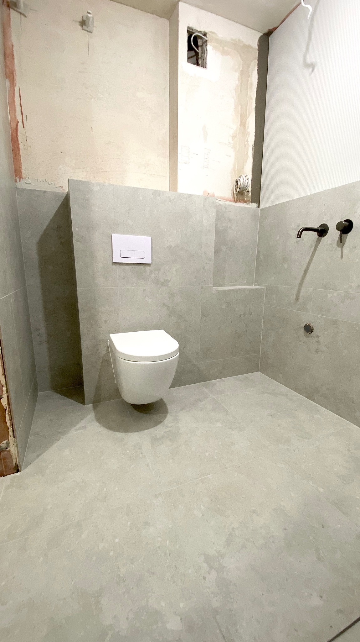 Szara łazienka z podwieszaną toaletą i czarną armaturą. Częściowo wykończone ściany, widoczne przewody. Minimalistyczny styl, surowe wykończenie.