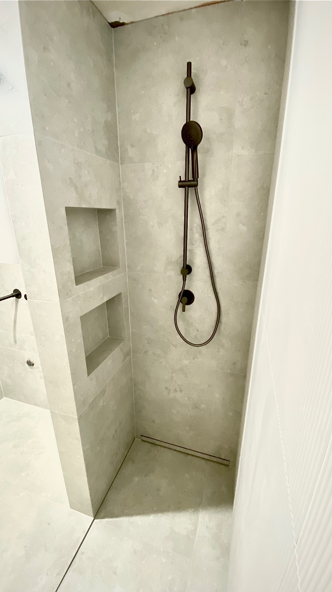 Szara łazienka z kabiną typu walk-in, wykończona płytkami imitującymi kamień. Widoczne wnęki w ścianie i czarna armatura.