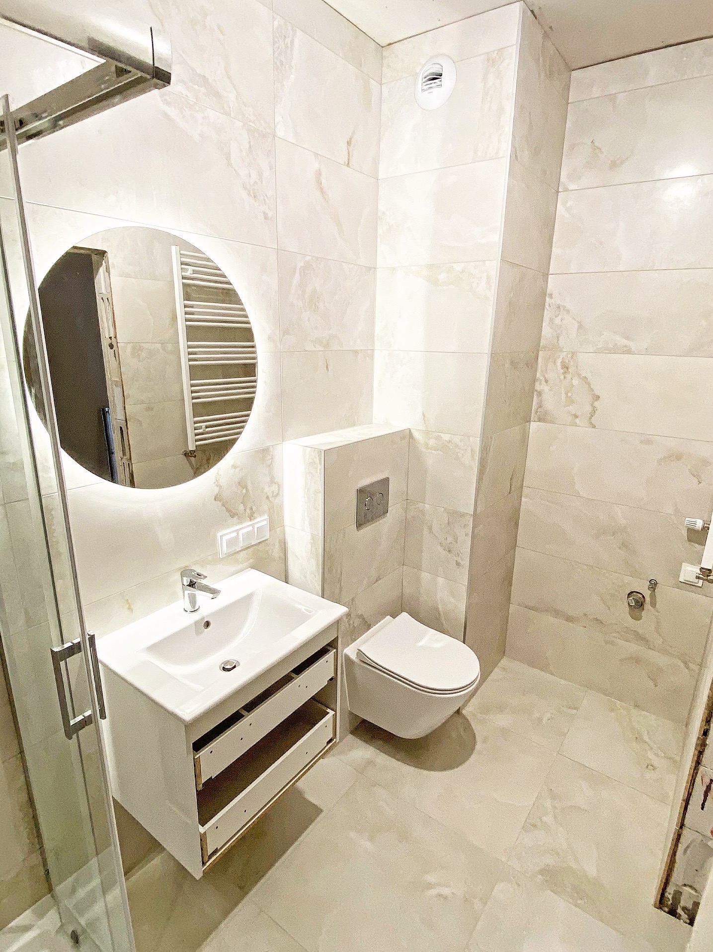 Jasna łazienka z beżowymi płytkami imitującymi marmur, okrągłe lustro z podświetleniem, wisząca toaleta i szafka pod umywalką z otwartymi szufladami.