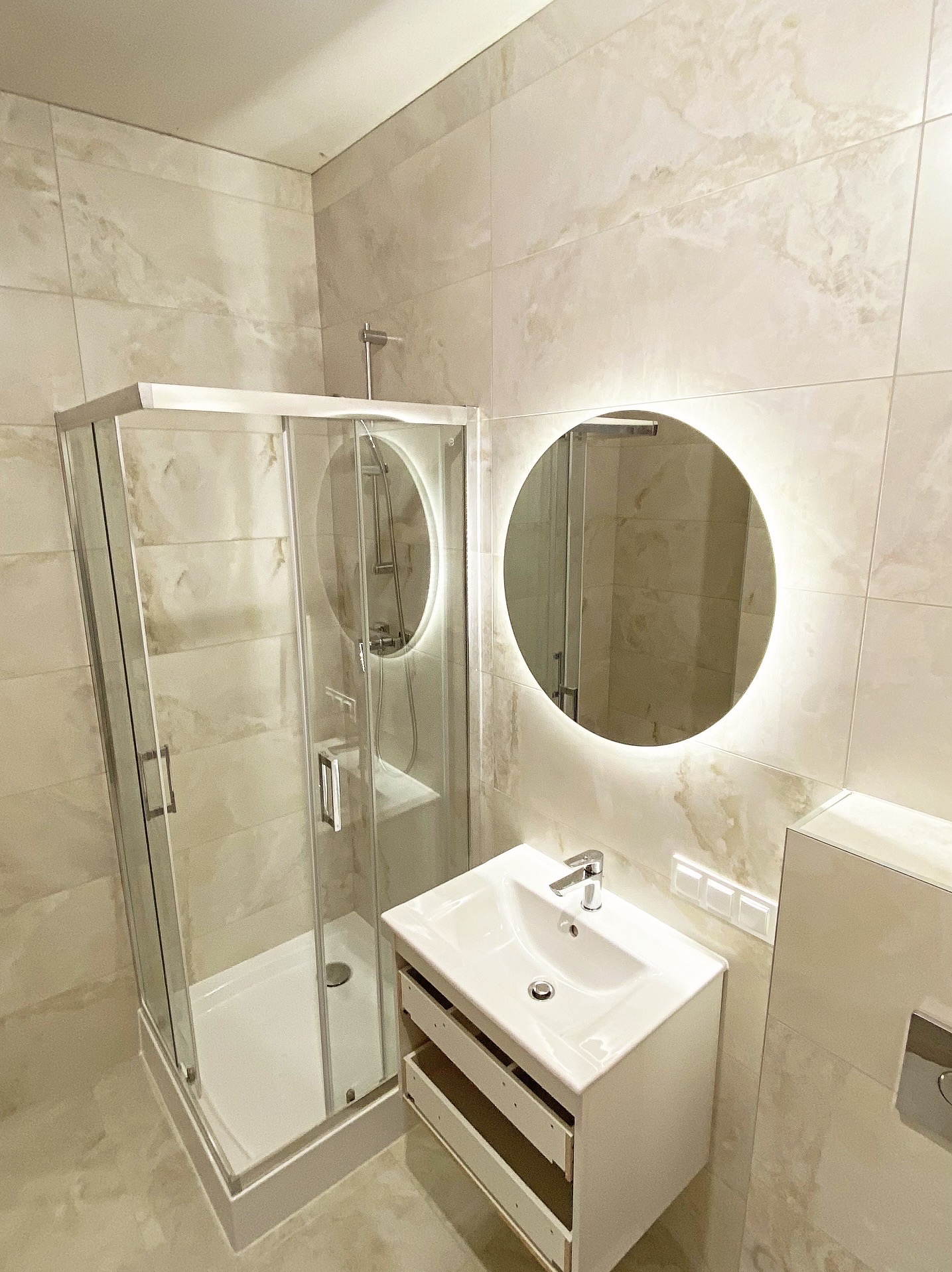 Wykończona łazienka z kabiną prysznicową, okrągłym lustrem z podświetleniem LED i białą szafką umywalkową z otwartymi szufladami. Beżowe płytki na ścianach i podłodze.