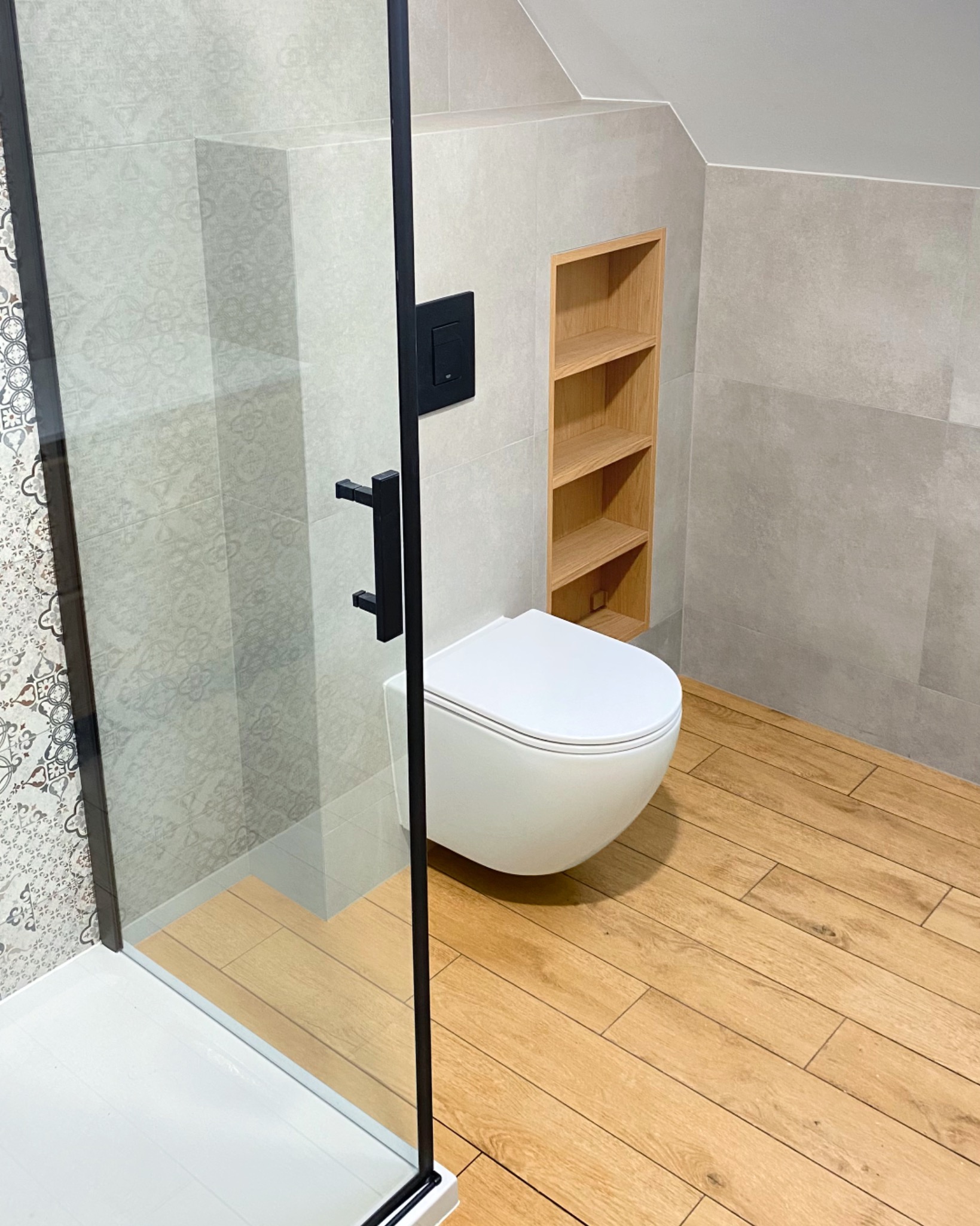 Nowoczesna łazienka z kabiną prysznicową typu walk-in, podwieszaną miską WC i drewnianą półką wnękową. Szare płytki i drewnopodobna podłoga tworzą spójną całość.
