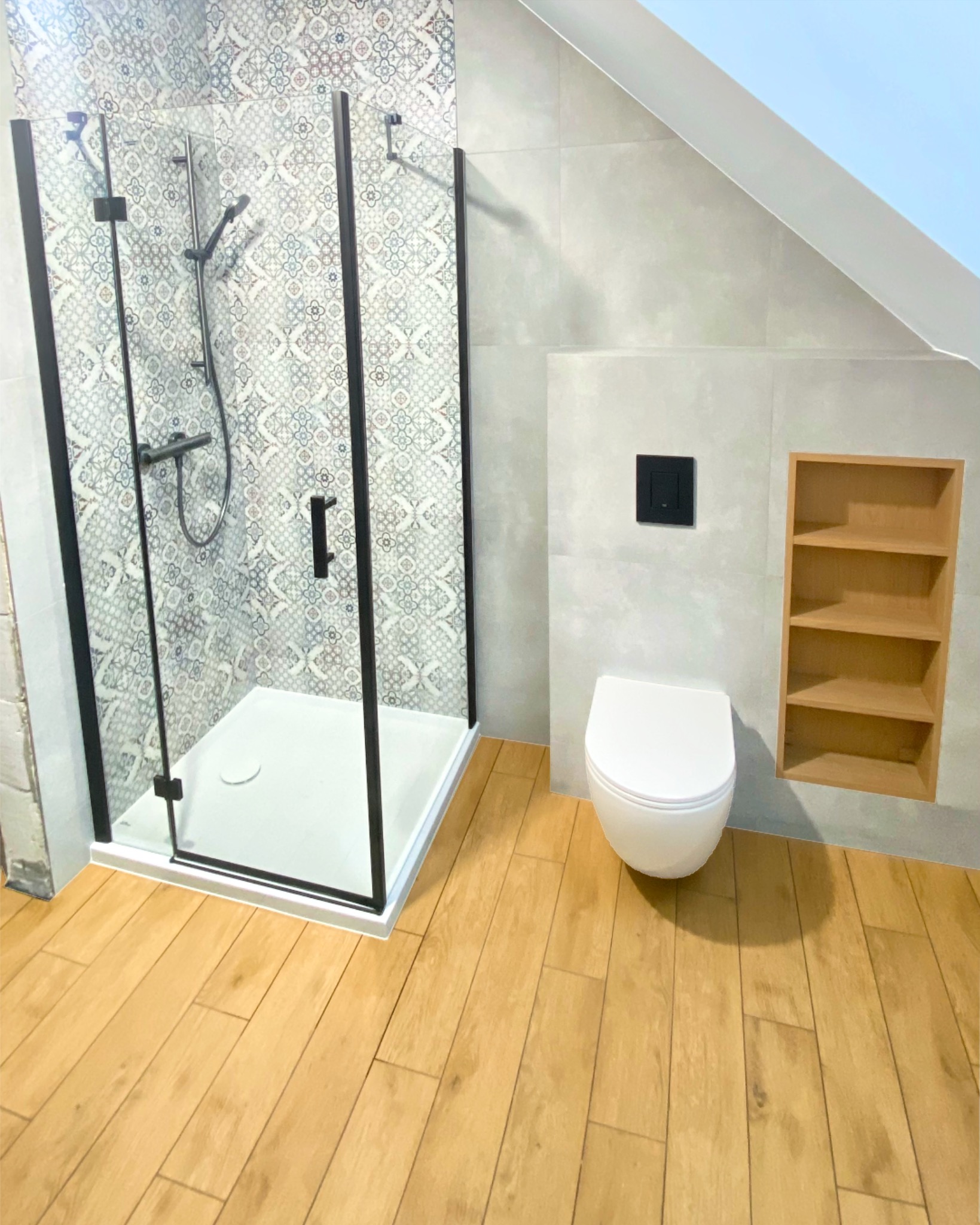 Nowoczesna łazienka na poddaszu: kabina prysznicowa z czarnymi profilami, toaleta wisząca i drewniane półki wnękowe. Płytki w geometryczne wzory i drewnopodobna podłoga.