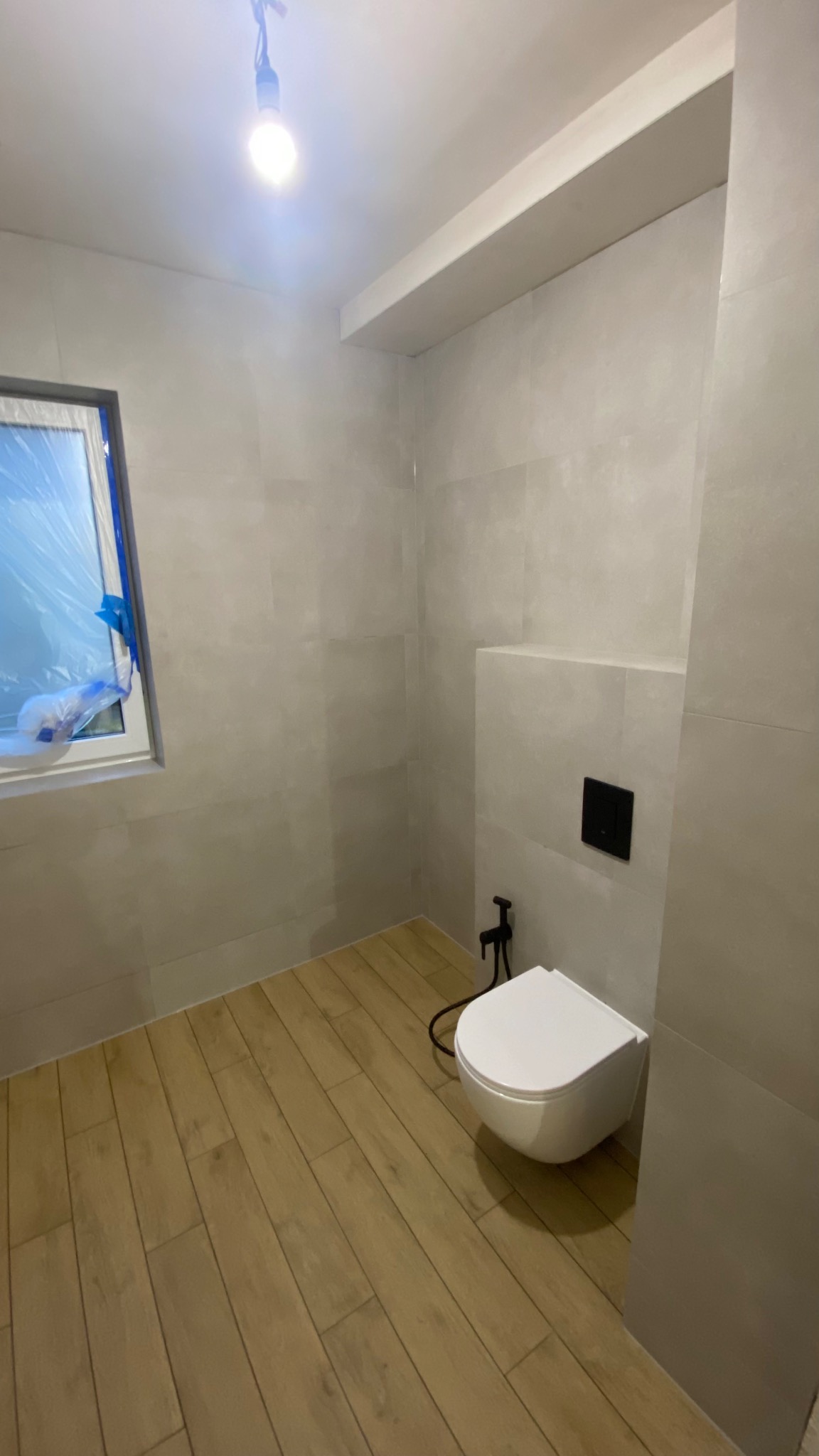 Jasna łazienka po remoncie w Warszawie: drewnopodobna podłoga, szare płytki ścienne, wisząca toaleta z czarną armaturą. Okno zabezpieczone folią.