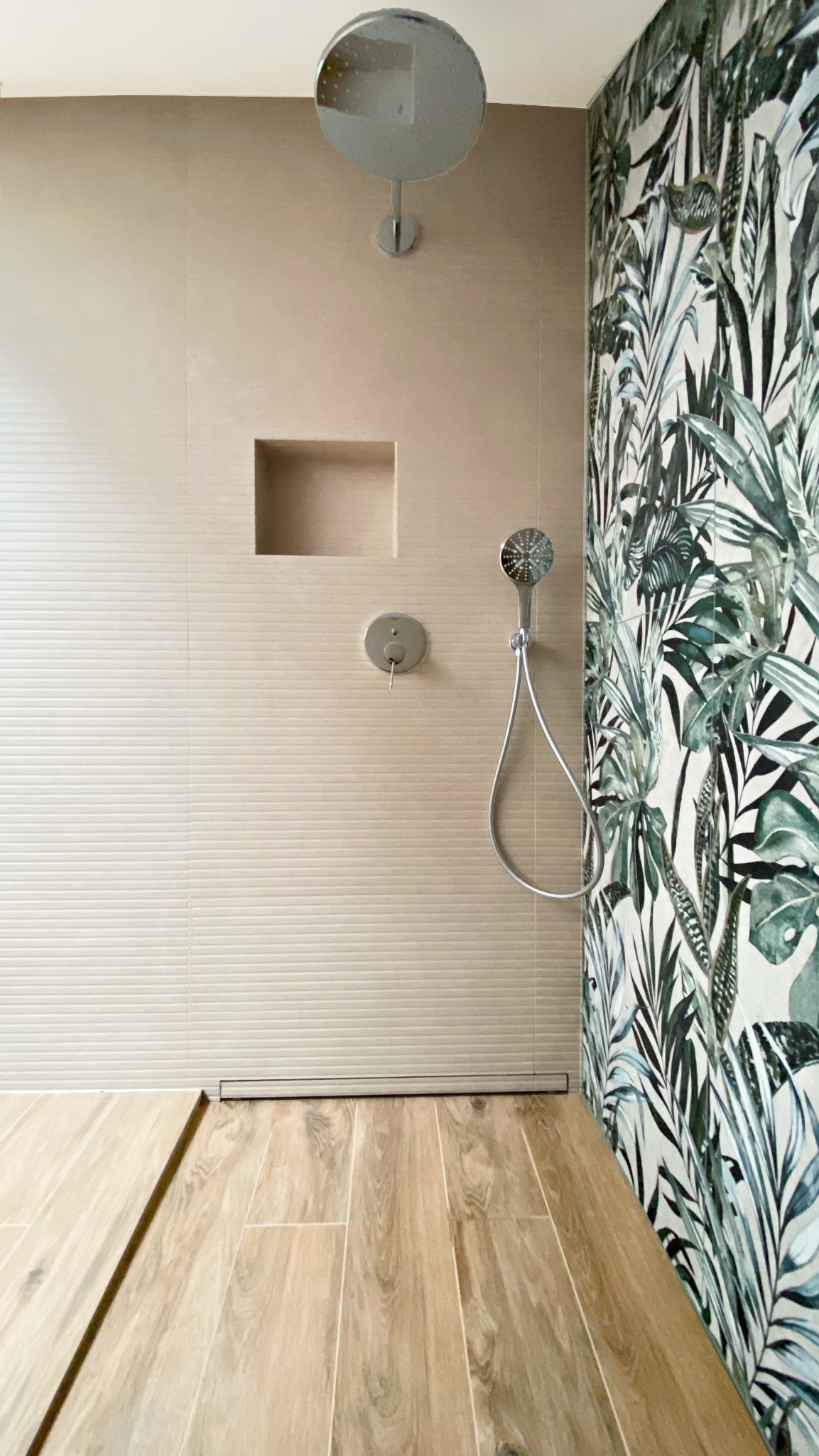 Nowoczesna kabina prysznicowa z beżowymi płytkami i akcentem tropikalnej tapety. Widoczna deszczownica, bateria i odpływ liniowy. Drewnopodobna podłoga.
