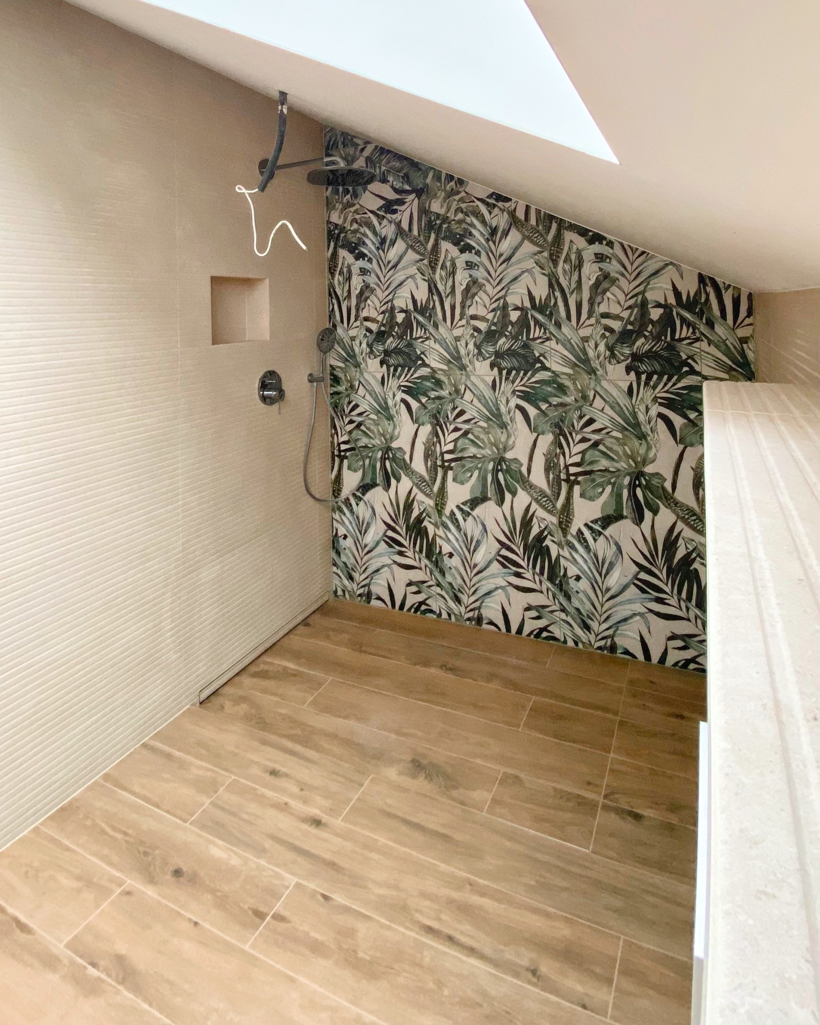 Wykończona łazienka na poddaszu z beżowymi płytkami imitującymi drewno na podłodze i ścianach, oraz ścianą akcentową w tropikalne liście. Widoczny prysznic.