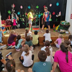 Iluzjonista w kolorowej koszuli prezentuje trik z linami na scenie przed grupą dzieci siedzących na podłodze. Na scenie dekoracje z kwiatów i drzew, skrzynie i kartony.