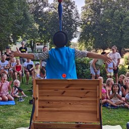 Art of Mind Norbert Ogoniak - Osoba w kostiumie z cylindrem i niebieską peleryną prezentuje drewnianą skrzynię przed grupą dzieci siedzących na trawie w parku.