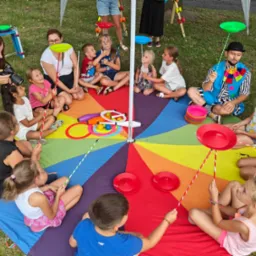 Grupa dzieci siedzi na kolorowej macie w kształcie koła, ucząc się kręcić talerzami na patyku pod okiem animatora w stroju klauna i osoby robiącej zdjęcia.