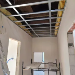 Montaż konstrukcji sufitu podwieszanego z ociepleniem wełną mineralną w Dobrzenicach, widoczne profile metalowe i fragment drewnianej konstrukcji dachu powyżej.