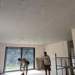 Zielony & JaRa - Montaż płyt gipsowo-kartonowych na suficie: dwóch mężczyzn pracuje w jasnym, przestronnym pokoju z dużym oknem, widoczne płyty GK zamocowane do sufitu, kable elektryczne zwisające z otworów...