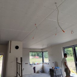 Zielony & JaRa - Wnętrze w trakcie prac wykończeniowych: zamontowany sufit z płyt gipsowo-kartonowych z widocznymi przewodami elektrycznymi, ściany w stanie surowym, okna z widokiem na zewnątrz, drabina oparta...