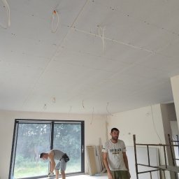 Zielony & JaRa - Montaż płyt karton gips na suficie w nowym budynku, widoczne kable elektryczne wystające z sufitu, pracownicy w trakcie pracy, duże okno w tle.