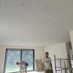 Montaż płyt karton gips na suficie w nowym budynku, widoczne kable elektryczne wystające z sufitu, pracownicy w trakcie pracy, duże okno w tle.