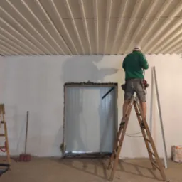 Pracownik na drabinie montuje listwę przy suficie z paneli, widoczna surowa ściana i otwór drzwiowy w trakcie prac wykończeniowych.