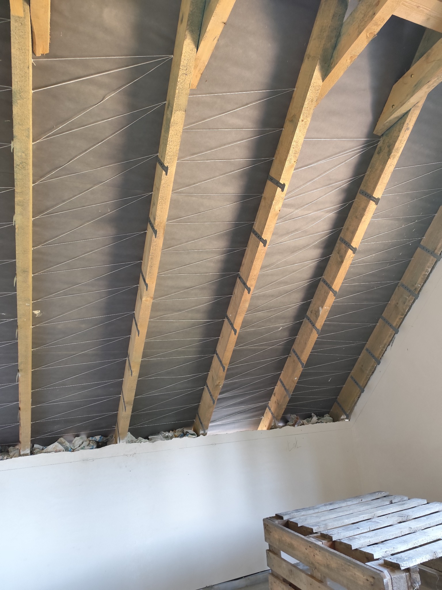 Poddasze w trakcie ocieplania: widoczne drewniane krokwie, szara folia paroizolacyjna z białymi sznurkami, częściowo wypełniona przestrzeń nad ścianą, oraz drewniana paleta w dolnym rogu.