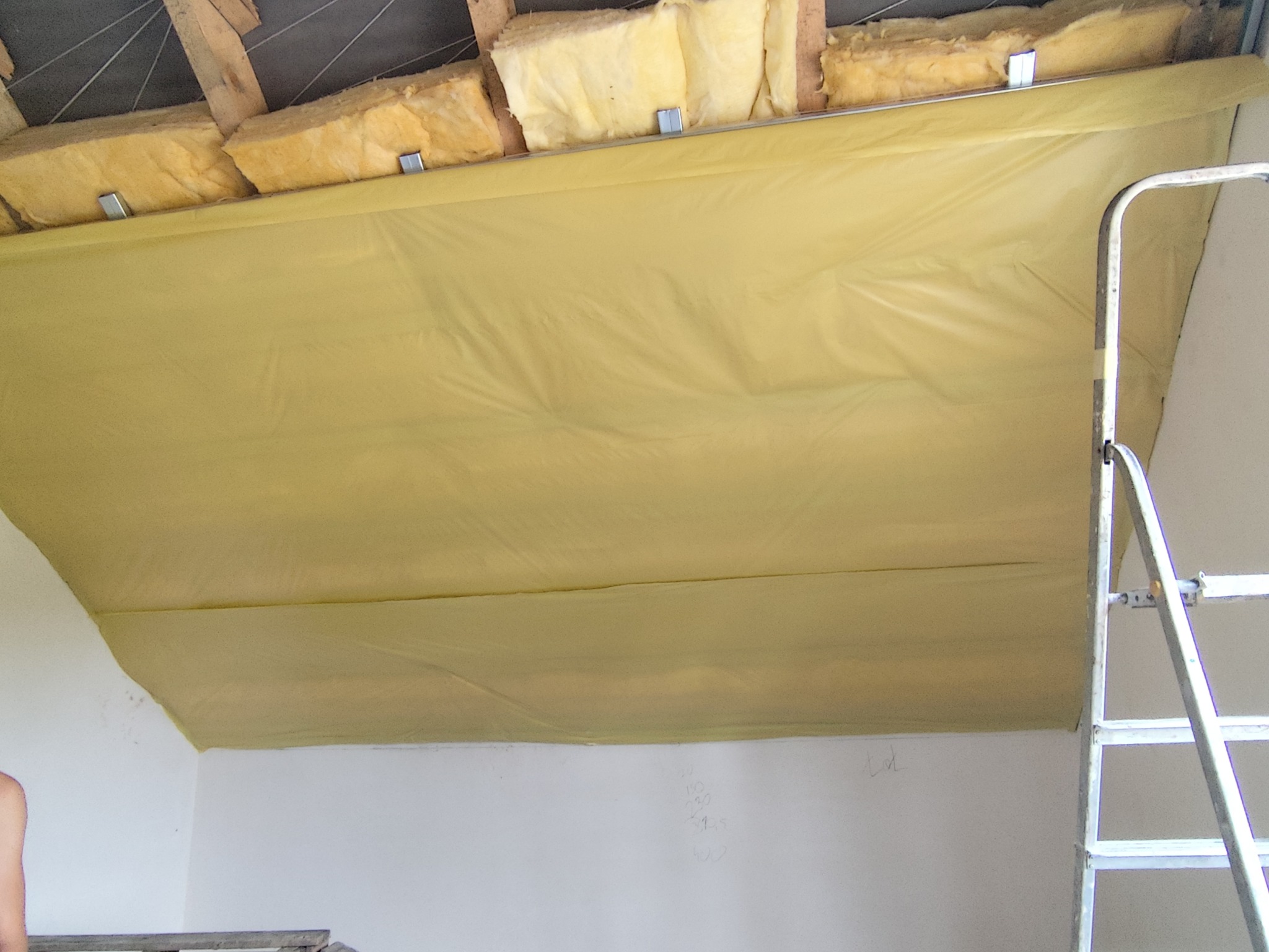 Ocieplenie poddasza wełną mineralną zabezpieczoną żółtą folią paroizolacyjną, widoczne elementy drewnianej konstrukcji dachu i drabina oparta o ścianę.