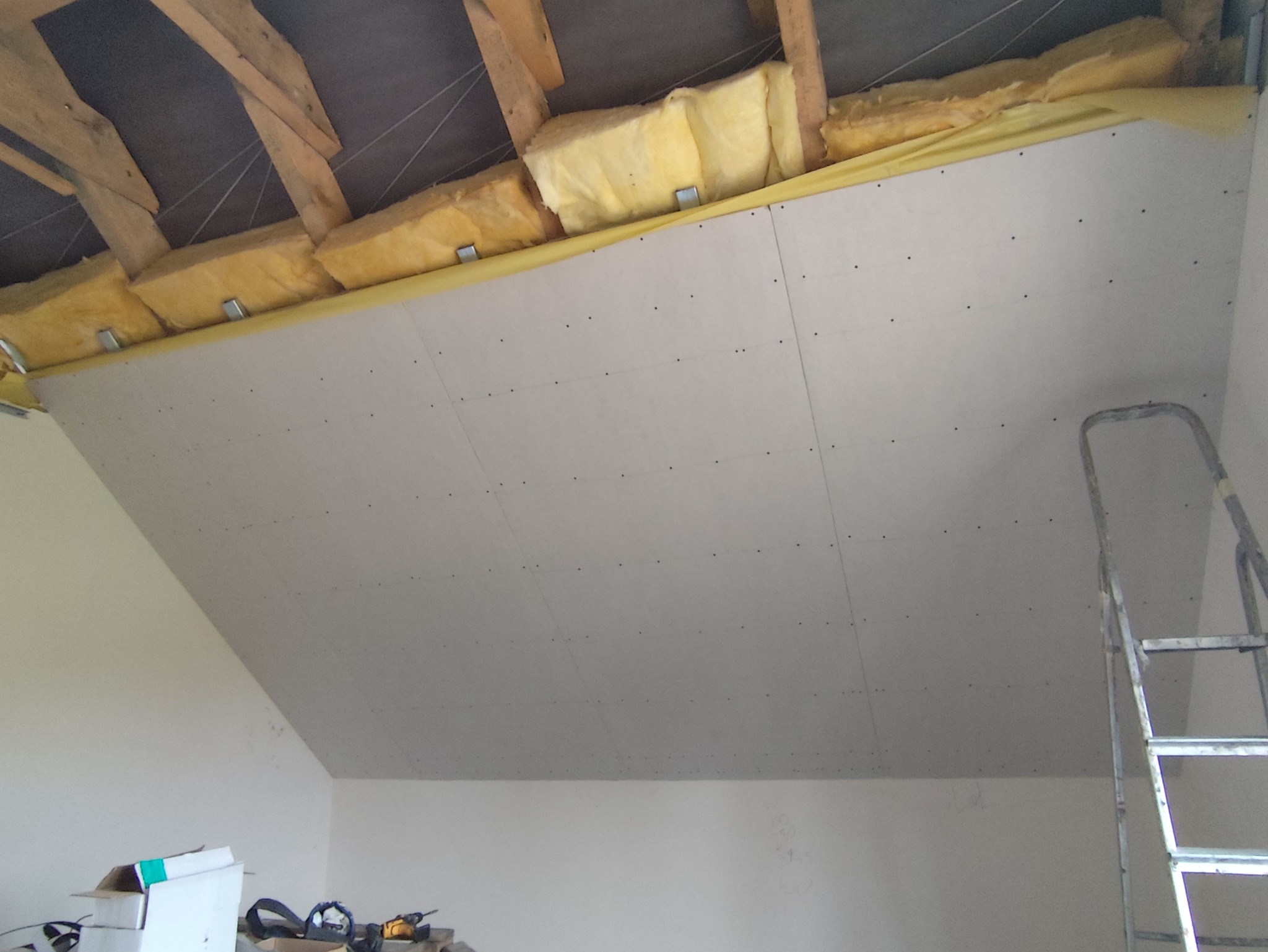 Poddasze z izolacją z wełny mineralnej i zamontowanymi płytami gipsowo-kartonowymi, widoczne drewniane elementy konstrukcyjne dachu i drabina oparta o ścianę.