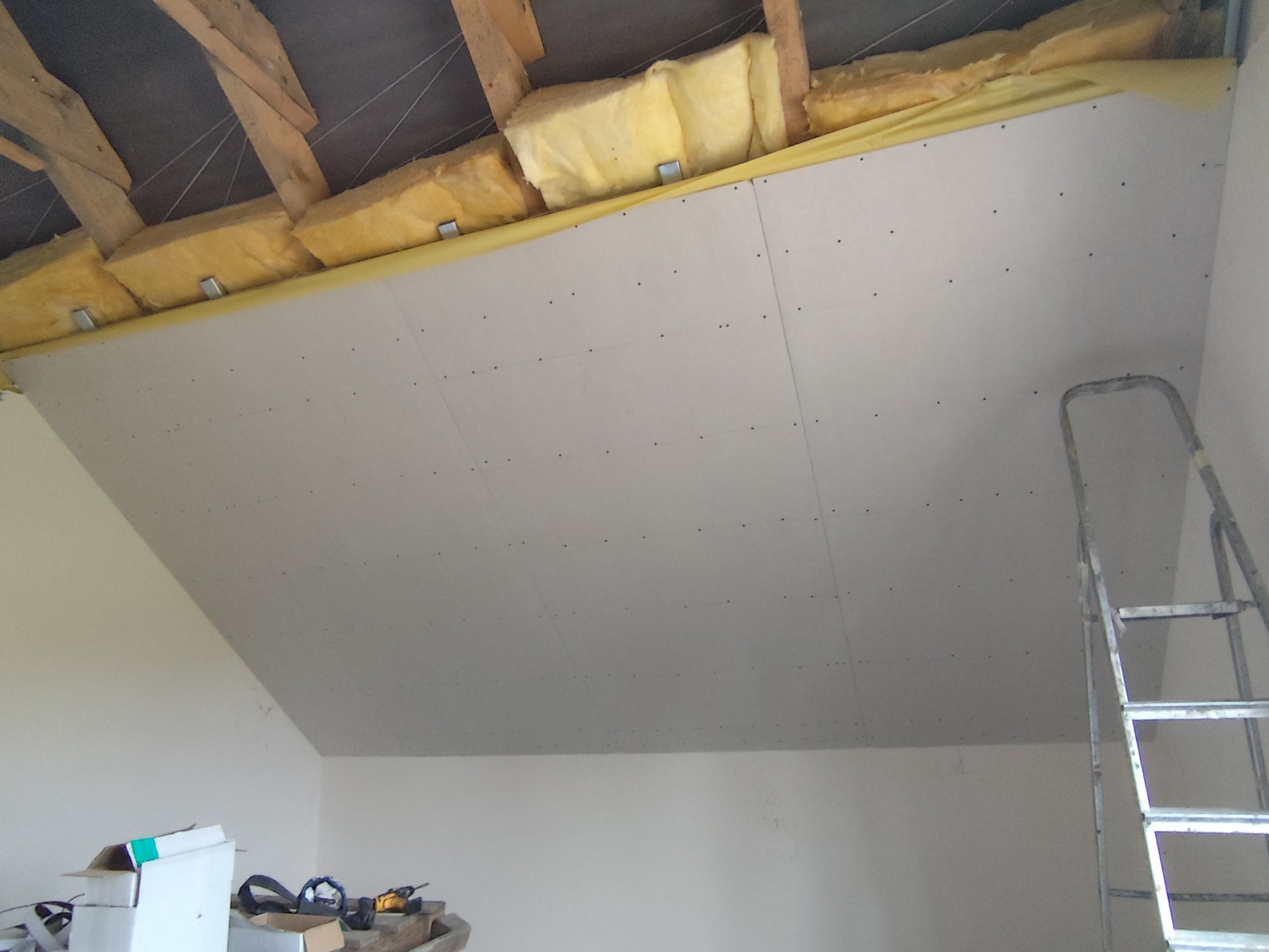 Poddasze z ociepleniem wełną mineralną, zabezpieczoną folią i zabudową z płyt gipsowo-kartonowych, widoczne ślady po wkrętach, drabina przystawiona do ściany, na podłodze narzędzia i kartony.