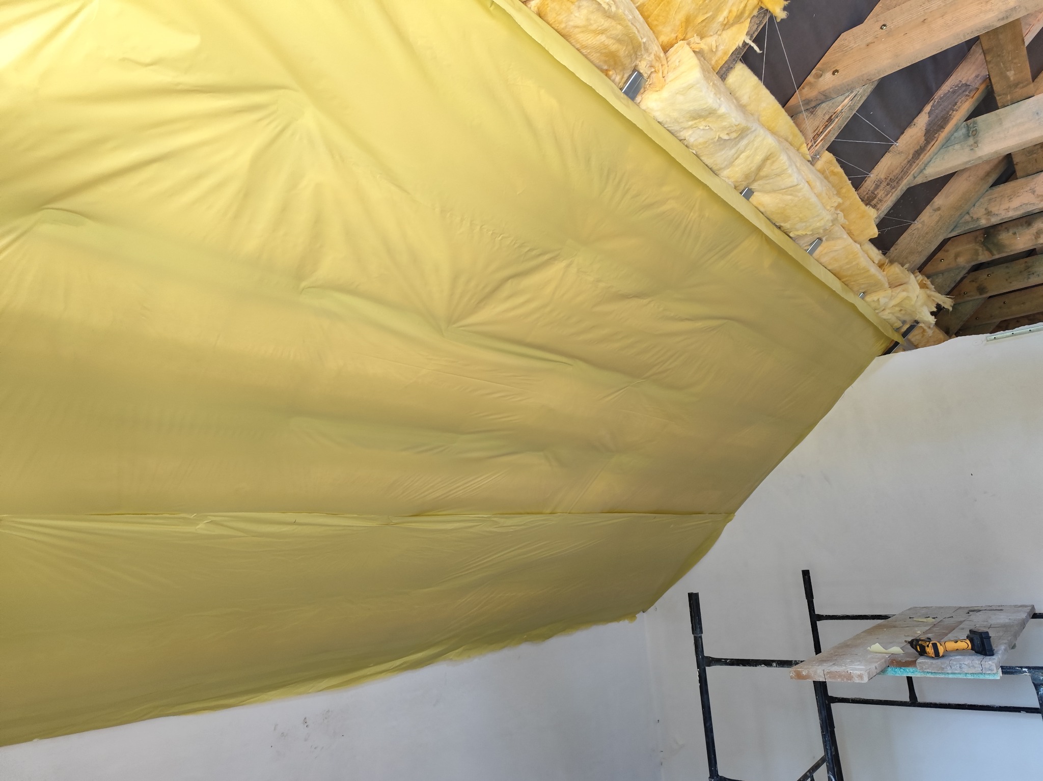 Izolacja poddasza wełną mineralną i folią paroizolacyjną, widoczne elementy więźby dachowej i rusztowanie z narzędziami.