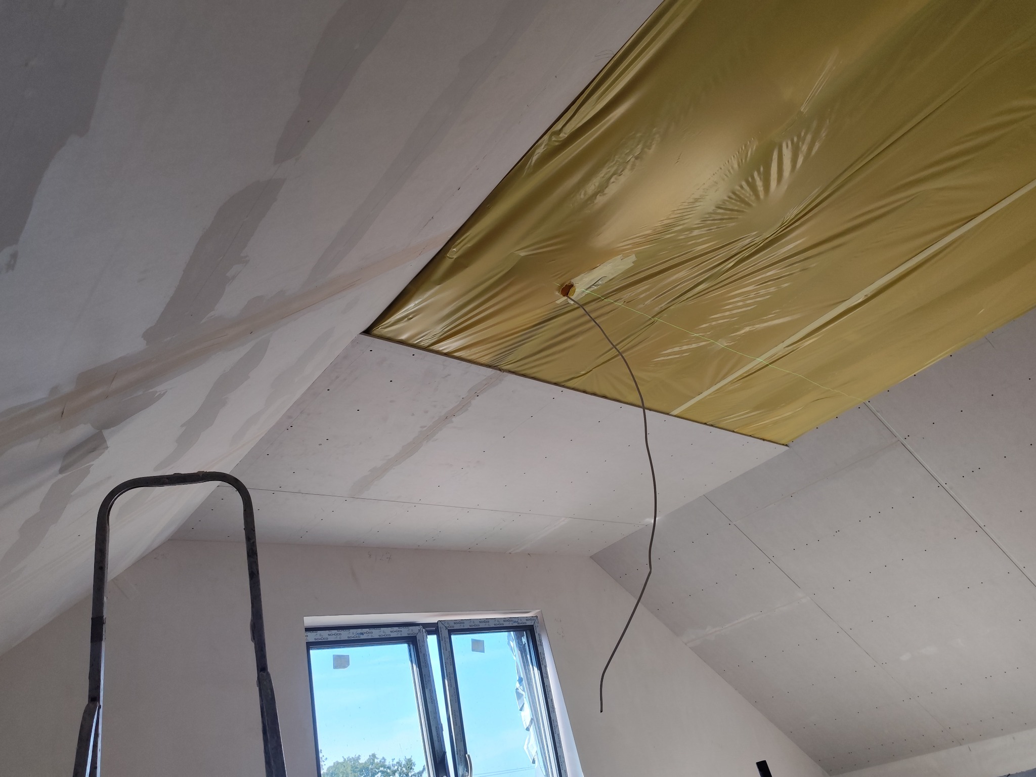 Montaż płyty karton gips w ocieplonym poddaszu, widoczna żółta folia zabezpieczająca izolację, częściowo zaszpachlowane łączenia płyt, drabina i okno dachowe w tle.