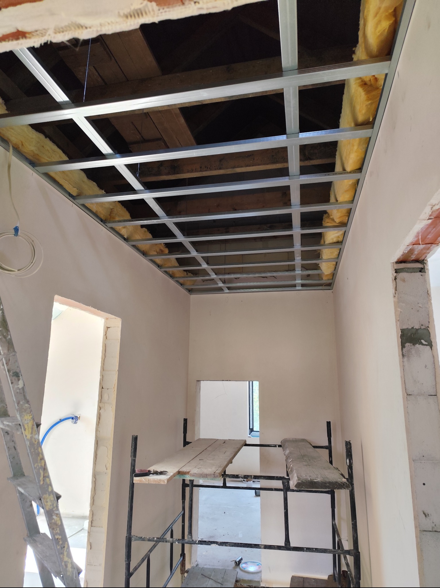 Montaż konstrukcji sufitu podwieszanego z ociepleniem wełną mineralną w Dobrzenicach, widoczne profile metalowe i fragment drewnianej konstrukcji dachu powyżej.