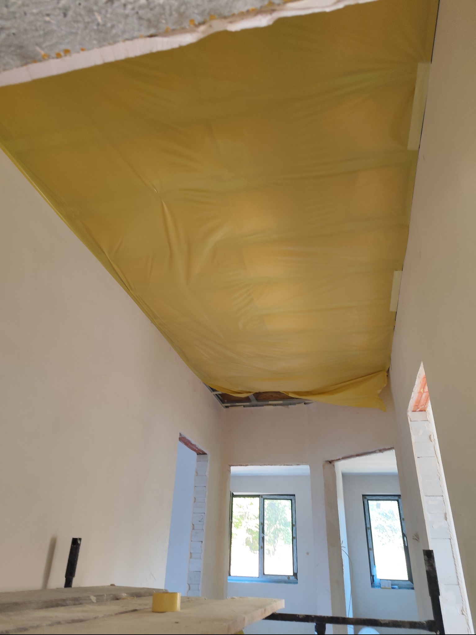 Żółta folia zabezpieczająca przymocowana taśmą do sufitu podczas prac wykończeniowych wnętrza, widoczne surowe ściany i przygotowane otwory drzwiowe.