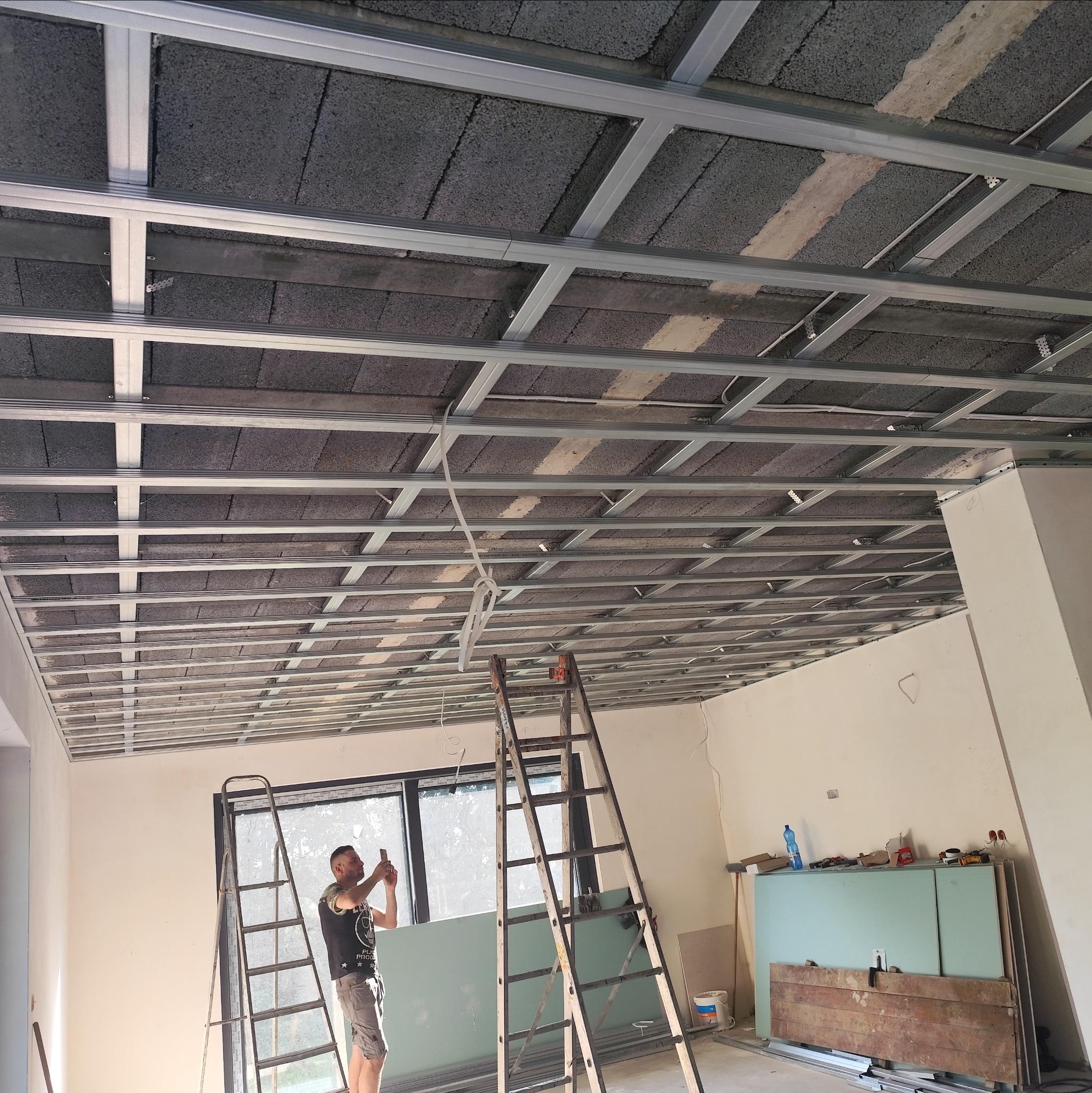 Montaż metalowej konstrukcji sufitu podwieszanego z widocznymi elementami izolacji, pracownik na drabinie wykonuje zdjęcie telefonem, w tle płyty gotowe do montażu.