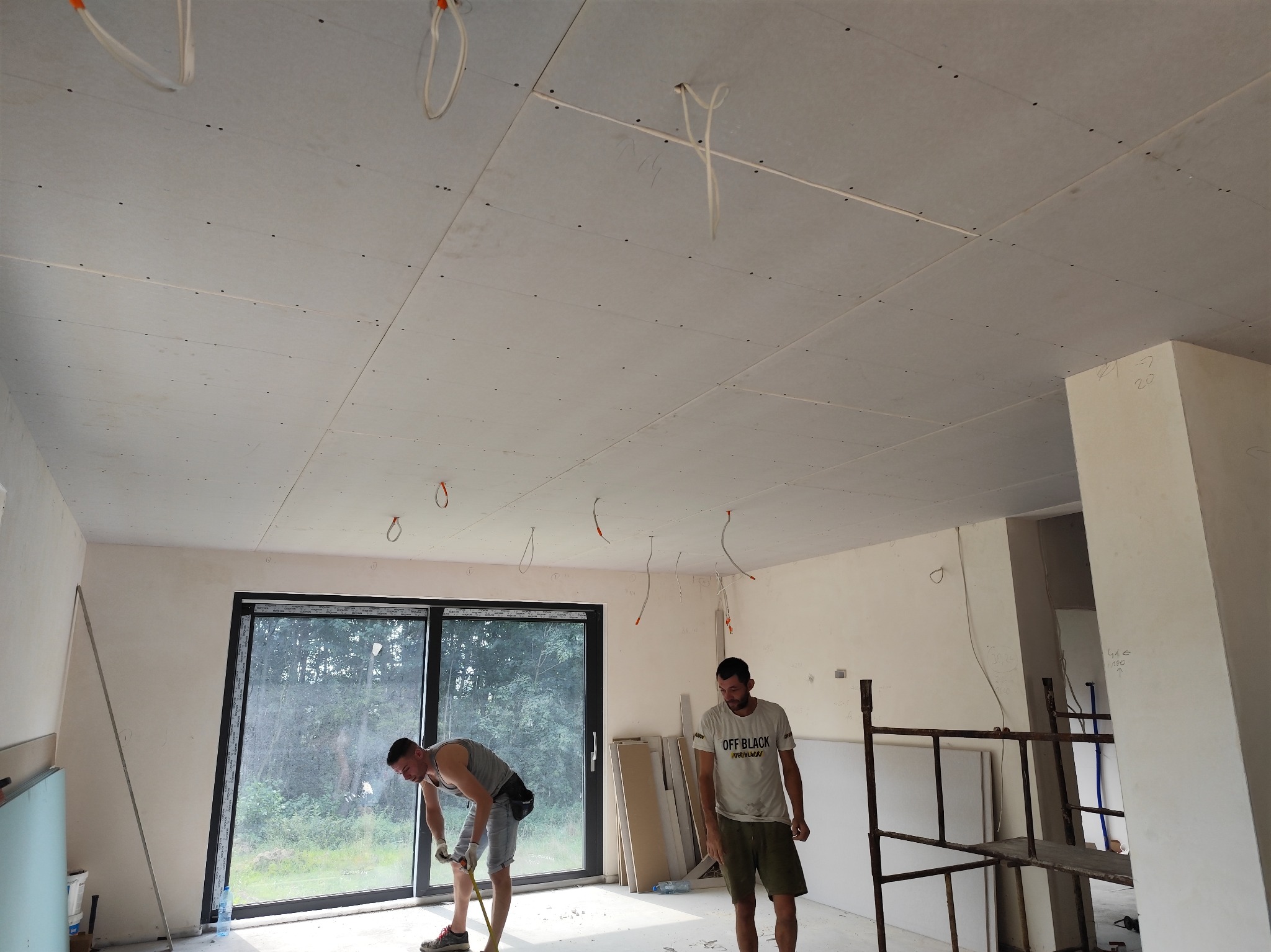 Montaż płyt gipsowo-kartonowych na suficie: dwóch mężczyzn pracuje w jasnym, przestronnym pokoju z dużym oknem, widoczne płyty GK zamocowane do sufitu, kable elektryczne zwisające z otworów...