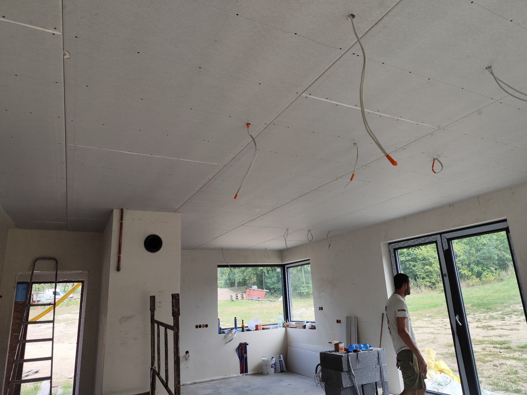 Wnętrze w trakcie prac wykończeniowych: zamontowany sufit z płyt gipsowo-kartonowych z widocznymi przewodami elektrycznymi, ściany w stanie surowym, okna z widokiem na zewnątrz, drabina oparta...