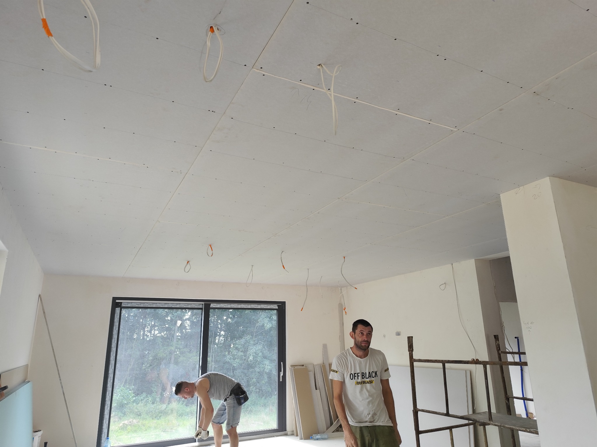Montaż płyt karton gips na suficie w nowym budynku, widoczne kable elektryczne wystające z sufitu, pracownicy w trakcie pracy, duże okno w tle.