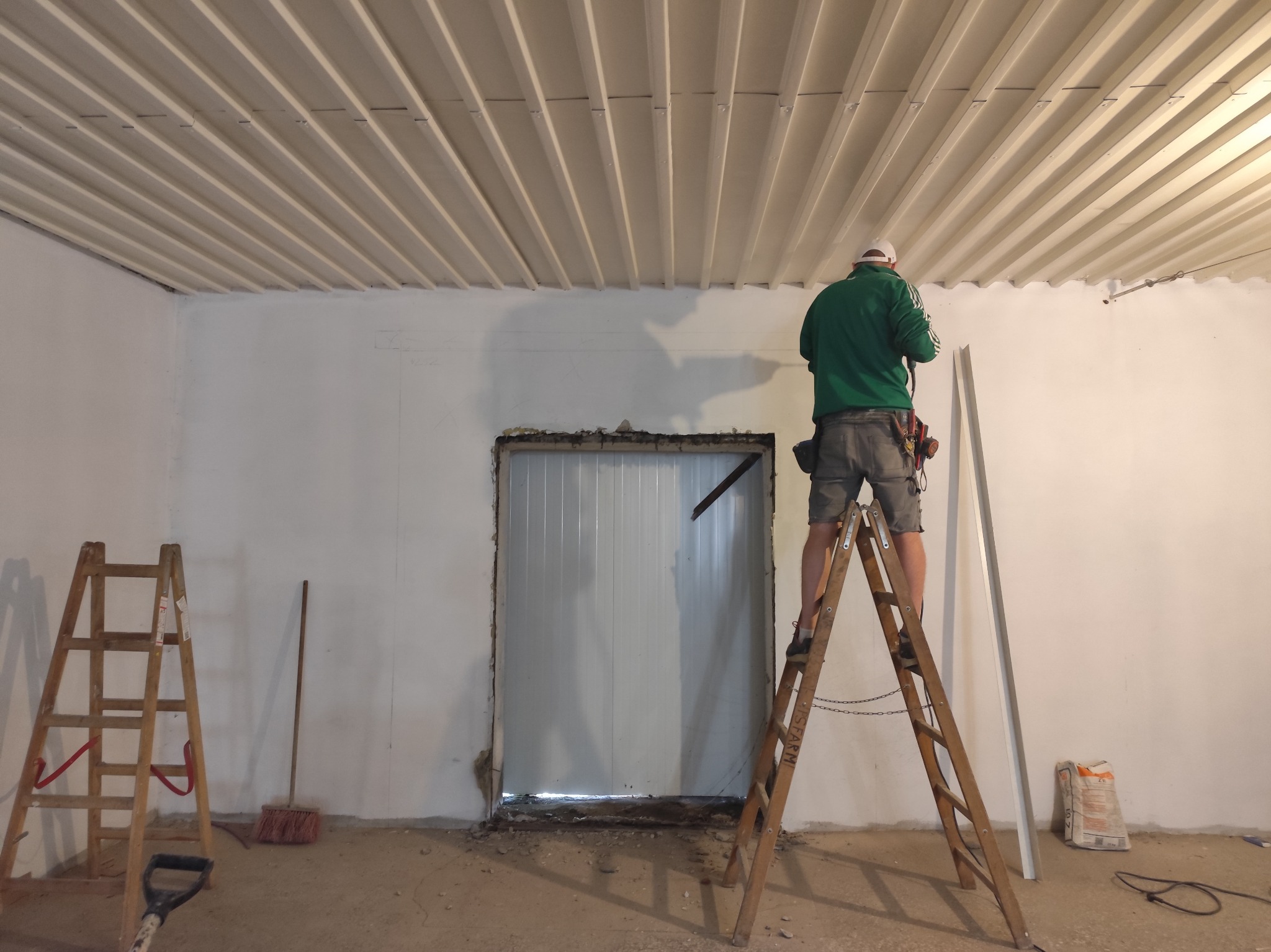 Pracownik na drabinie montuje listwę przy suficie z paneli, widoczna surowa ściana i otwór drzwiowy w trakcie prac wykończeniowych.