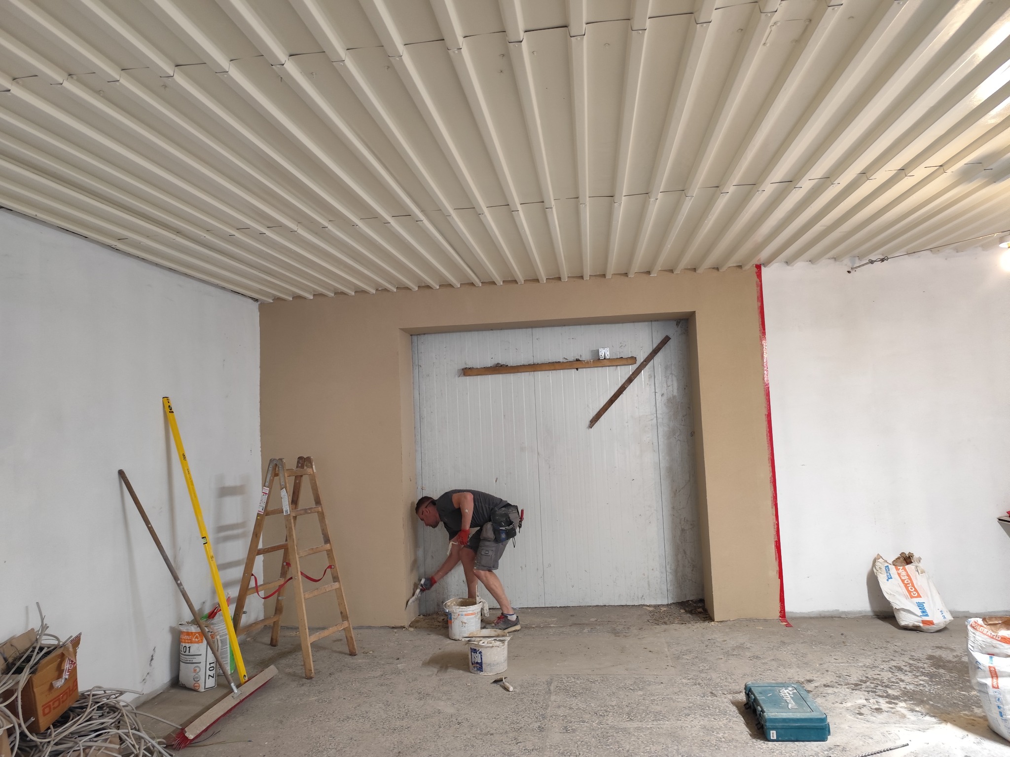 Pracownik w szarym t-shircie i krótkich spodenkach szpachluje ścianę w pomieszczeniu z metalowym sufitem, widoczne narzędzia, drabina i worki z zaprawą.