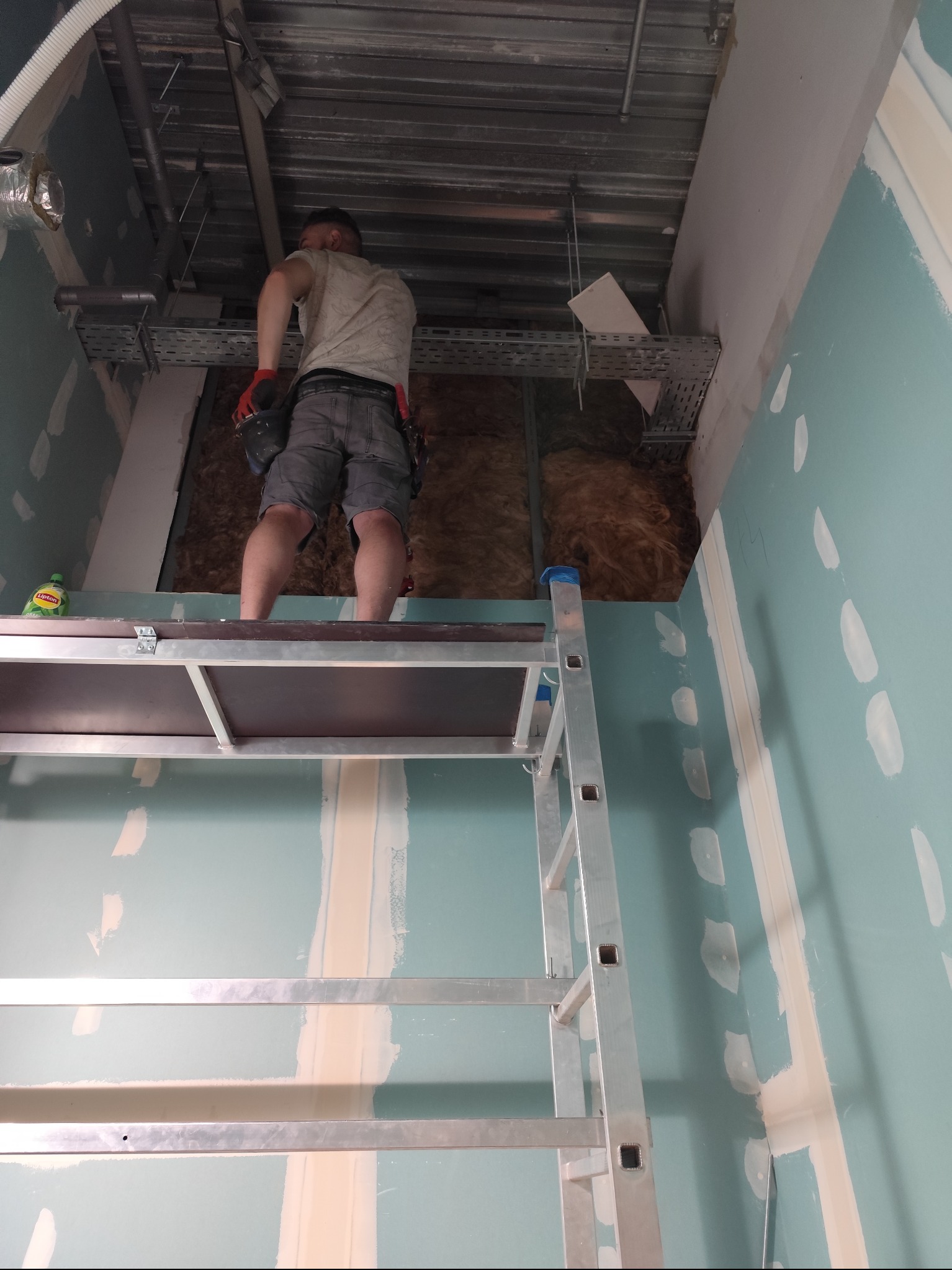 Pracownik na rusztowaniu montuje izolację poddasza, widoczne płyty gipsowo-kartonowe przed szpachlowaniem, butelka z napojem na rusztowaniu.