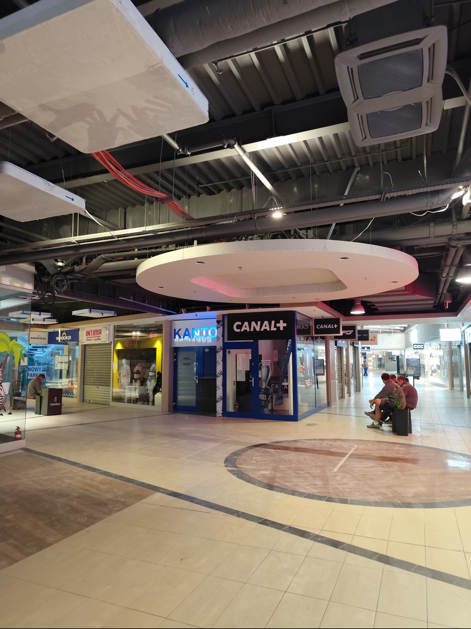 Wnętrze galerii handlowej z widocznymi sufitami podwieszanymi, instalacjami wentylacyjnymi i oświetleniem, oraz witrynami sklepów w tle.