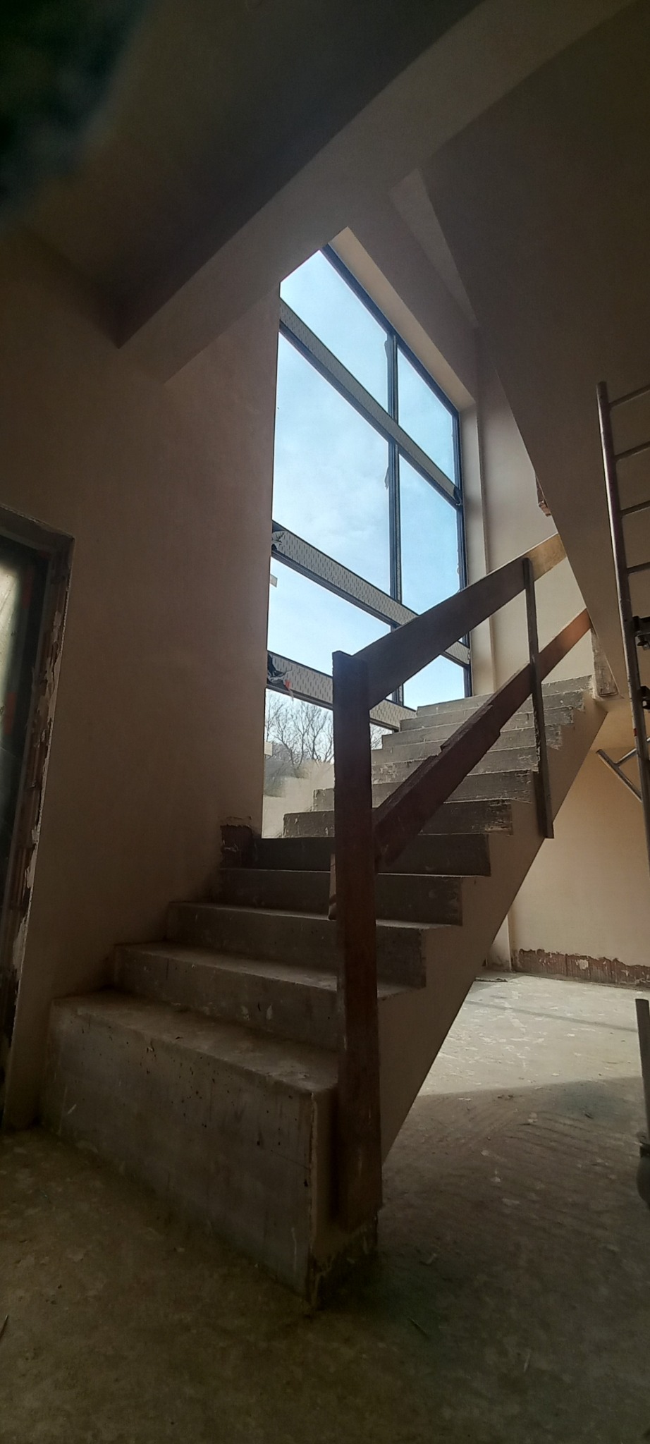 Surowe betonowe schody z drewnianą barierką w surowym stanie, widoczne ślady po tynku, duże okno z widokiem na błękitne niebo.