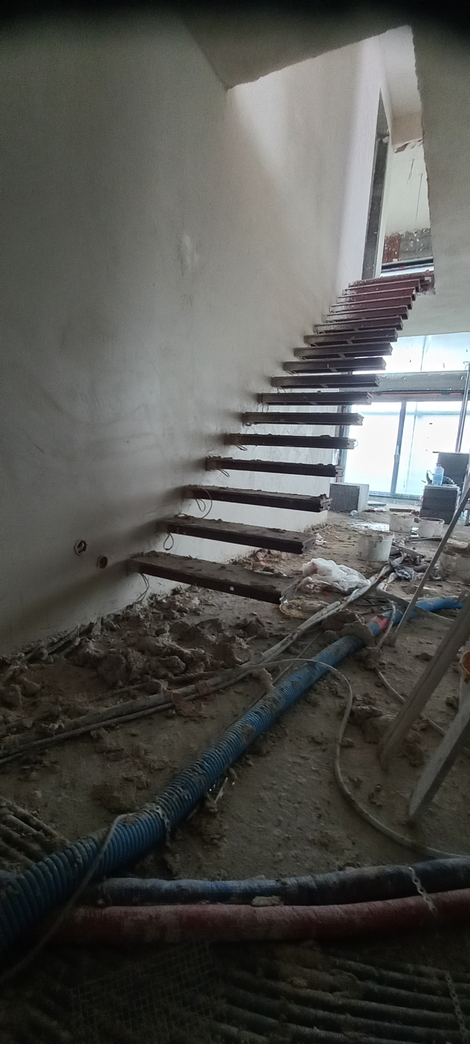 Świeżo otynkowana ściana przy schodach z widocznymi drewnianymi stopniami w trakcie remontu, niebieskie rury na podłodze i okno w tle.