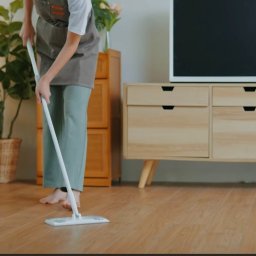 ELITE CLEAN Joanna Ziemiakowska - Osoba w fartuchu myje drewnianą podłogę mopem w jasnym salonie z roślinami doniczkowymi i komodą. W tle telewizor i sofa z narzutą.