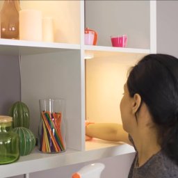 ELITE CLEAN Joanna Ziemiakowska - Osoba czyści półkę w białym regale z dekoracjami. Widoczna butelka ze środkiem czyszczącym i ściereczka. Oświetlenie półki podkreśla detale.