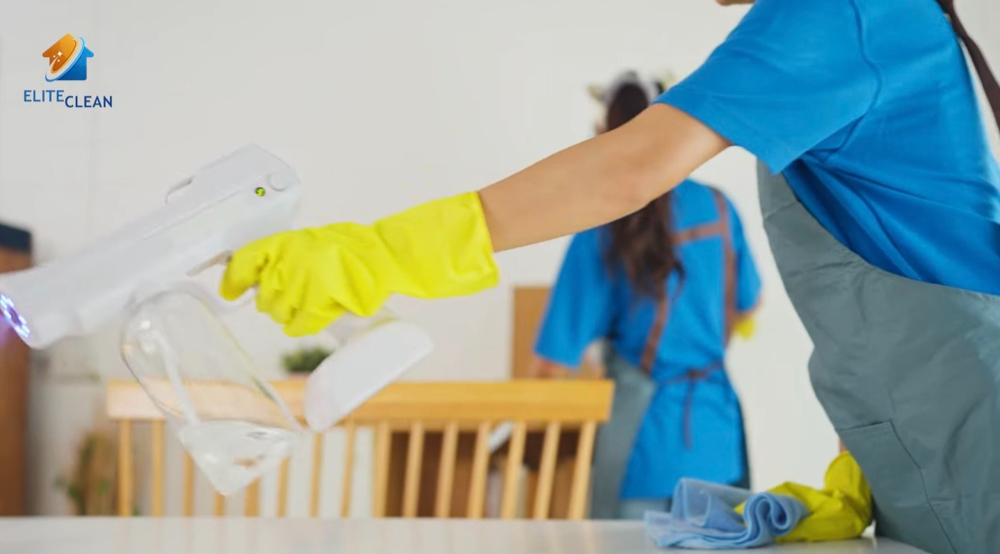 Pracownik w żółtych rękawicach dezynfekuje stół za pomocą białego pistoletu do dezynfekcji UV, w tle drugi pracownik. Nowoczesna metoda dezynfekcji.