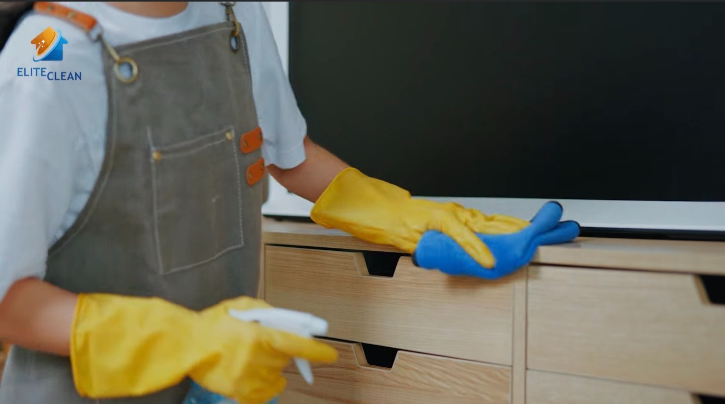 Sprzątanie szafki RTV: osoba w fartuchu i żółtych rękawicach czyści mebel niebieską szmatką i spryskiwaczem. Widoczne logo firmy Elite Clean.