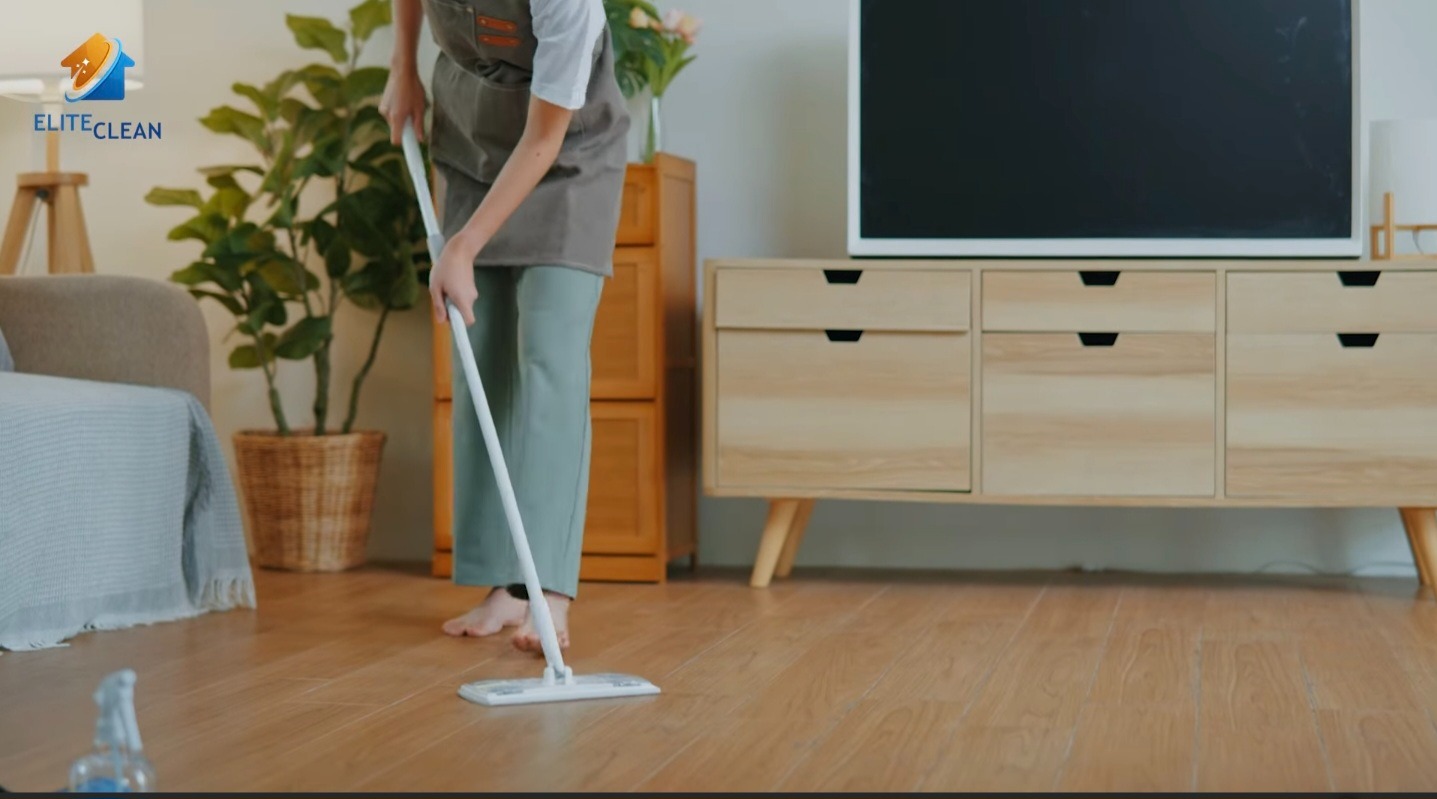Osoba w fartuchu myje drewnianą podłogę mopem w jasnym salonie z roślinami doniczkowymi i komodą. W tle telewizor i sofa z narzutą.