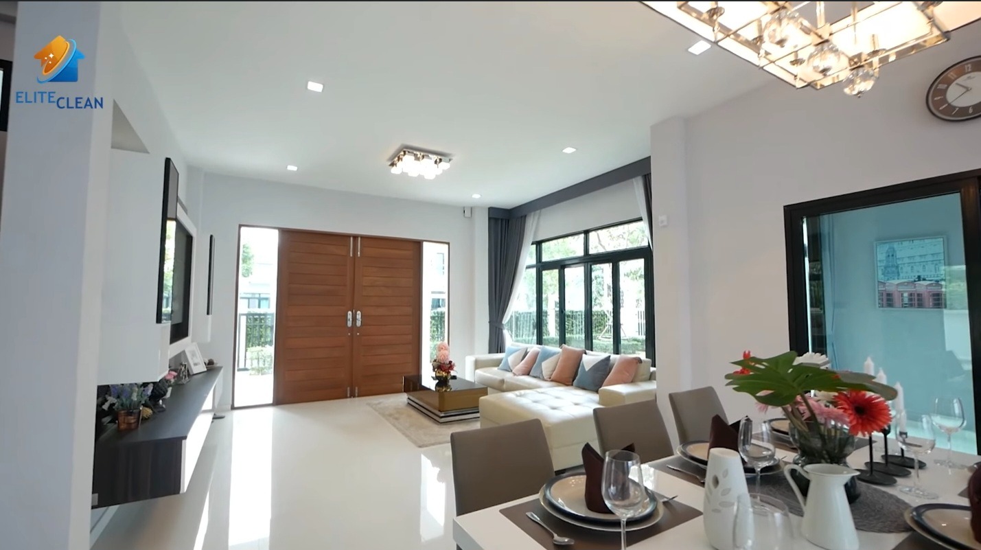 Jasne wnętrze domu po sprzątaniu: białe ściany i podłogi, stół z zastawą, sofa z poduszkami, drewniane drzwi. Widoczny znak firmy sprzątającej.