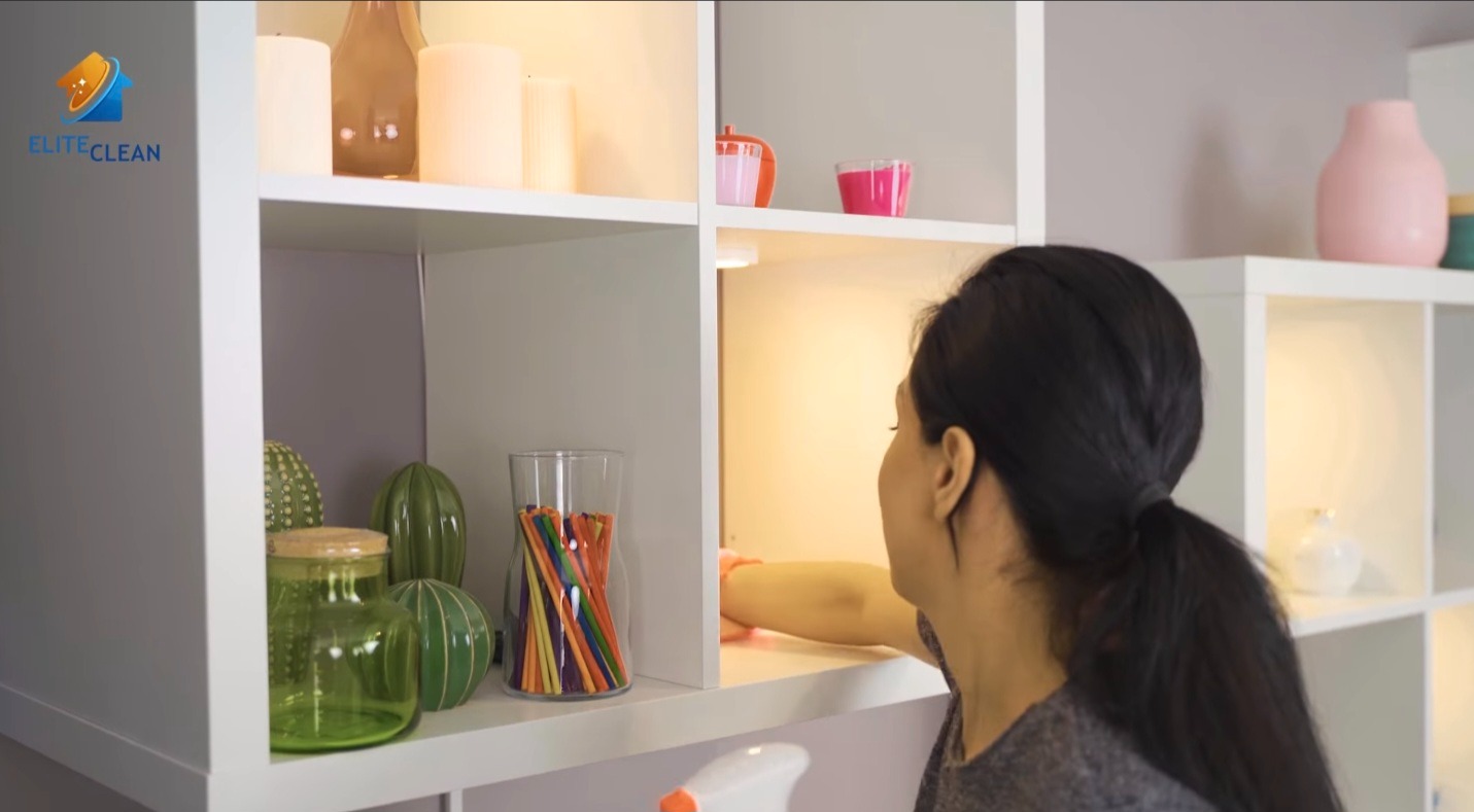 Osoba czyści półkę w białym regale z dekoracjami. Widoczna butelka ze środkiem czyszczącym i ściereczka. Oświetlenie półki podkreśla detale.