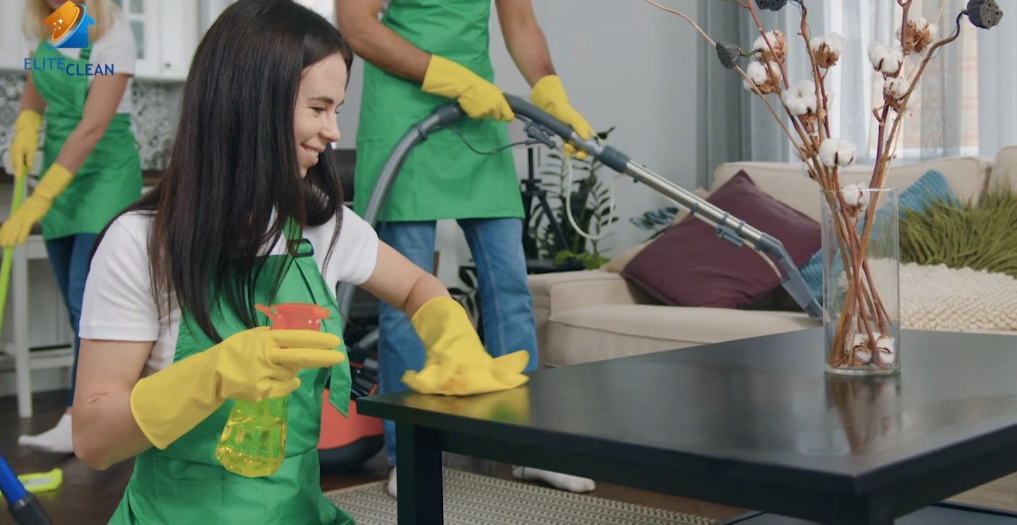 Zespół sprzątający w akcji: kobieta w żółtych rękawicach czyści stół sprayem, w tle odkurzacz i inni pracownicy. Wazon z dekoracją na stole.