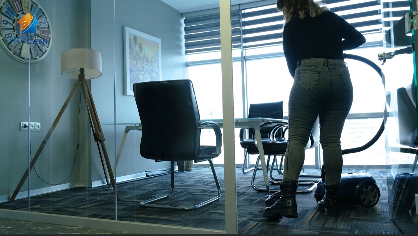 Pracownica sprzątająca biuro odkurzaczem. Widoczne biurko, krzesła, lampa na trójnogu i zegar z logo firmy sprzątającej. Duże okno z żaluzjami w tle.